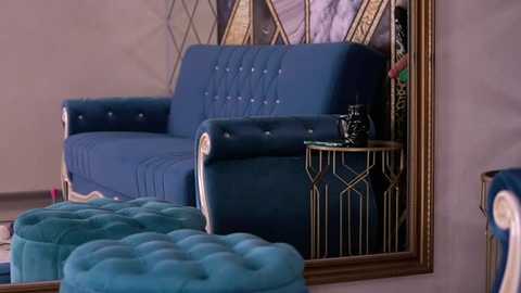 Media: Video of a chic living room featuring a mid-century modern blue velvet sofa with tufted detailing and gold accents, a blue tufted ottoman, and a black and gold side table with a black vase. The background includes a large, ornate gold-framed mirror and a decorative screen.