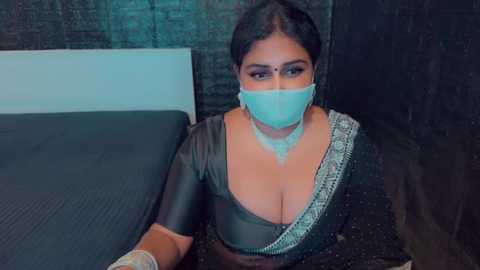 Media: Video of a South Asian woman with dark hair, wearing a green blouse, gold necklace, and a face mask, lying on a bed with a black quilt, in a dimly lit room with a dark brick wall.