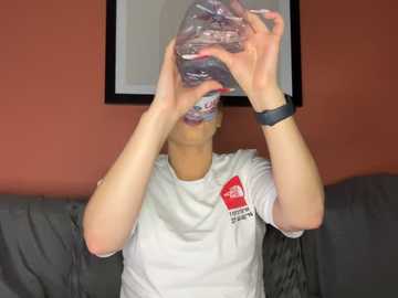 Media: A video of a light-skinned person, wearing a white T-shirt and black watch, drinking from a clear plastic bottle, with a red logo on the shirt, in a room with a grey couch and orange walls.