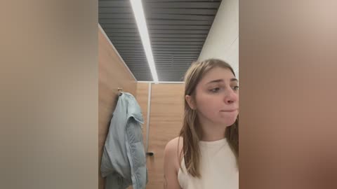 Media: Video of a young woman with straight, light brown hair in a white tank top, standing in a narrow, modern bathroom with beige walls, wooden paneling, and a gray ceiling.