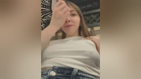 Media: Video of a young woman with fair skin and brown hair, wearing a white tank top and high-waisted blue jeans, taking a selfie in a dimly lit room with a black wall and white patterned tapestry.