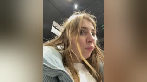 Media: A video of a young woman with long, wet hair, wearing a light blue jacket, taken in a dimly lit room with fluorescent lighting and blurred, indistinct background.