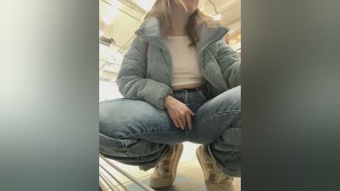 Media: Video of a young woman crouching in a bathroom. She wears a light blue puffer jacket, high-waisted blue jeans, and tan boots. The background features a mirror, white tiles, and a shower curtain.