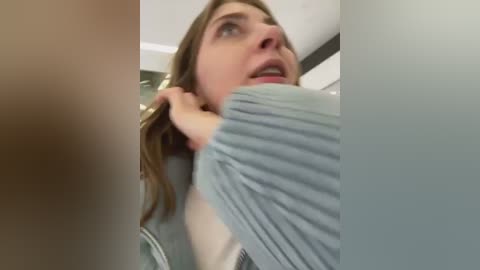 Media: Video of a Caucasian woman with long brown hair, wearing a grey sweater, pulling a grey shirt over her head. Background is blurred, suggesting an indoor setting.