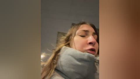 Media: Video of a young woman with light skin, long brown hair, and a light blue hoodie, looking distressed, possibly in pain, in a dimly lit room with beige walls.