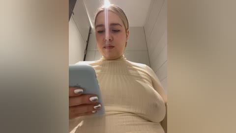 Media: Video of a plus-sized woman with blonde hair, wearing a tight, cream-colored ribbed turtleneck, holding a smartphone, in a dimly lit bathroom with white tiled walls.