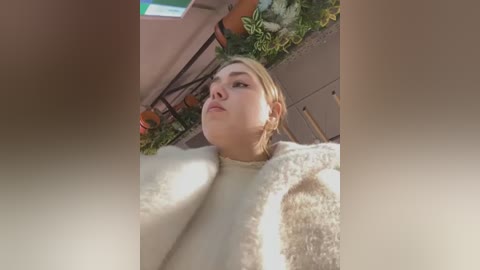 Media: Video of a blonde woman in a cozy, white, fluffy sweater, standing in a greenhouse filled with potted plants and hanging baskets.