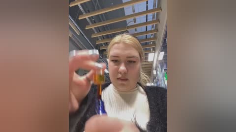 Media: Video of a young woman with light skin and blonde hair, wearing a white turtleneck and dark cardigan, holding a small glass bottle with a yellow liquid. Background shows a modern, industrial space with wooden beams and fluorescent lighting.