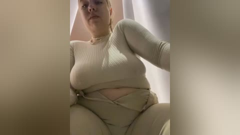 Media: A video of a plus-sized woman with light skin, wearing a tight beige ribbed top and matching high-waisted pants, seated indoors with light-colored curtains in the background.