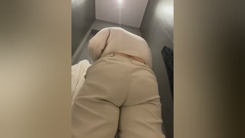 Media: Video of a person lying facedown on a bed in a small, dimly lit room. The person wears beige pants and a white shirt, their buttocks prominently visible. The walls are grey, and a light bulb hangs from the ceiling.