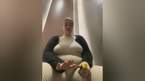 Media: A video of a plus-size woman in a beige jumpsuit and dark sweater sitting in a narrow, beige-tiled hallway, holding a banana. She has light skin and is looking down at her hands.