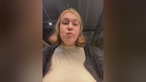 Media: Video of a young woman with light skin and shoulder-length blonde hair, wearing a beige turtleneck and a dark blue cardigan, making a pouty face in a car.