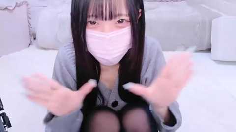 Media: A video of an Asian woman with long black hair, wearing a pink mask, sitting on a white bed, making a hand gesture, in a minimalist, white bedroom.