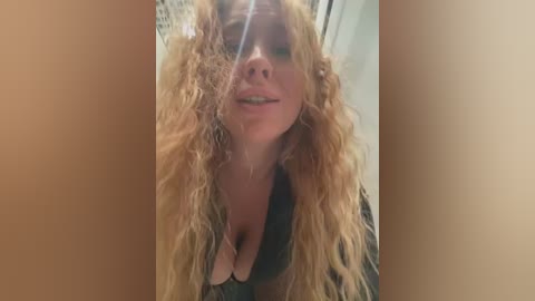 Media: Video of a woman with long, curly blonde hair, wearing a black top, smiling with eyes closed. Background is blurred, indoors, likely bathroom setting.