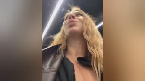 Media: Video of a blonde woman in a black leather jacket, looking upward with a slight smile, taken in a dimly lit, modern setting with fluorescent lights.