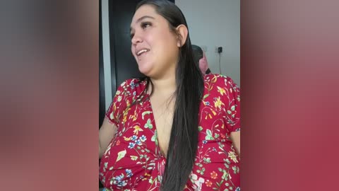 Media: Video of a Latina woman with long, straight black hair, wearing a red floral dress with a plunging neckline, smiling, indoors with blurred background.