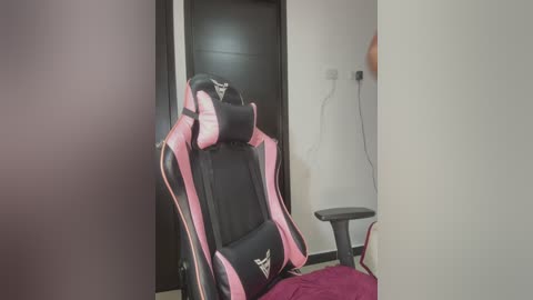 A video of a pink and black gaming chair with a white logo, in a modern, minimalist room with white walls, a black door, and a red chair.