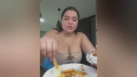 A video shows a plus-sized woman with dark hair, wearing a strapless top, eating fries and dipping them in a yellow sauce.