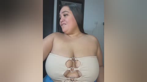 Video of a plus-sized woman with long black hair, wearing a beige, strapless top with cut-out details, seated indoors against a white wall.