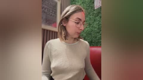 Media: Video of a young Caucasian woman with light brown hair and glasses, wearing a light-colored sweater, seated in a red-leather booth, with a green wall and framed artwork in the background.