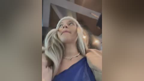 Video of a blonde woman with long hair in a deep blue one-shoulder dress, gazing upward, indoors with wooden beams and soft lighting.