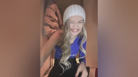 Video of a young woman with long, wavy blonde hair, wearing a white beanie and blue long-sleeved top, laughing and leaning against a brown quilted jacket.