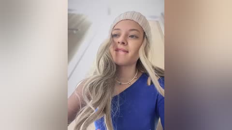 Video of a young woman with long, wavy blonde hair, wearing a beige beanie and a blue sweater. She has a light skin tone and a confident smile. Background is blurry.