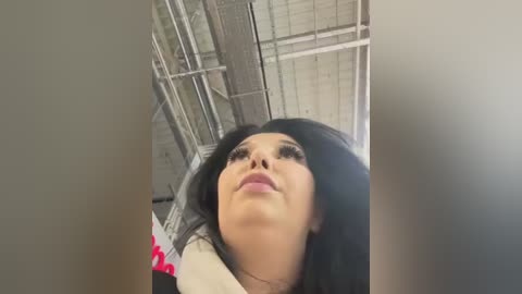 Media: A close-up video of a woman with long black hair and light skin, wearing heavy makeup, including dark eyeliner and mascara. She gazes upwards. The background shows a metal ceiling and a blurry red object.