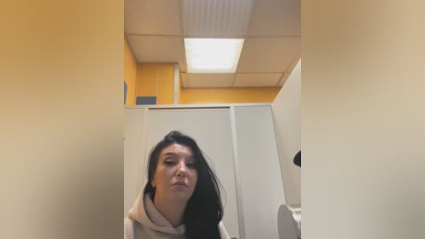 Media: Video of a young woman with long black hair, wearing a beige hoodie, standing in a yellow-walled restroom with white cabinets and a fluorescent ceiling light.