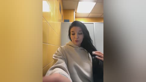 Media: Video of a young woman with long black hair, wearing a beige hoodie, standing in a yellow-tiled bathroom with a white shower stall, a fluorescent light overhead, and a blurred person in the background.