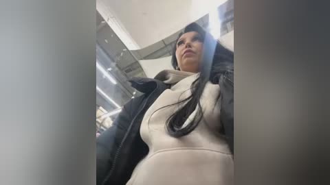 Media: Video of a woman with long black hair, wearing a black leather jacket over a white turtleneck sweater, looking down a subway station's escalator.