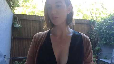 Media: A video of a young Caucasian woman with shoulder-length brown hair, wearing a low-cut black top and open brown cardigan, standing outdoors in a garden with a wooden fence and greenery in the background.