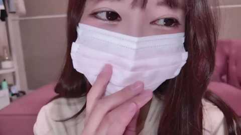 Media: Video of an Asian woman with long brown hair and light skin, wearing a white surgical mask, touching her mouth with her fingers, in a pink room.