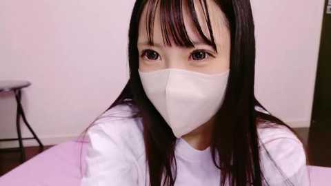 Media: Video of an Asian woman with long black hair and bangs, wearing a white surgical mask, white medical gown, and sitting on a pink surface in a sterile, white-walled room.