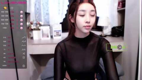 Media: A young East Asian woman with fair skin, long brown hair, and small breasts sits in a dimly lit room, wearing a sheer black top, with a blurred background and a digital camera interface overlay.