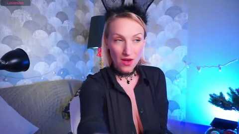 Media: Video of a blonde woman in black bunny ears, wearing a low-cut black shirt, seated in a room with white floral wallpaper, a black lamp, and Christmas decorations.