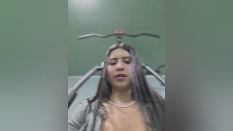 Media: Video of a young woman with long brown hair, wearing a gray cardigan, lying on a metal rack with her head held by straps, in an industrial setting.