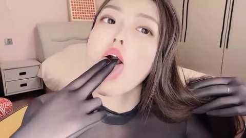 Media: A video of a young woman with fair skin and long brown hair, wearing a sheer black top, having her mouth licked by black-gloved hands in a softly lit, pink-themed bedroom.