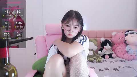 Media: Video of an East Asian woman with glasses, wearing a white top, sitting on a pink gaming chair in a cozy bedroom with plush toys.