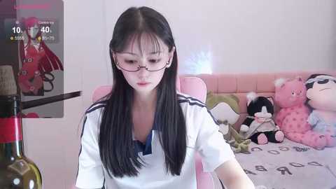 Media: Video of an Asian woman with long black hair and glasses, wearing a white polo shirt, sitting at a pink gaming chair in a brightly lit bedroom.