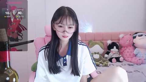 Media: A video of an Asian woman with long black hair and glasses, wearing a white polo shirt, sitting on a pink couch with stuffed animals.