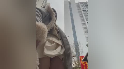 Media: Video of a woman wearing a gray cardigan and white skirt, walking on a city street with tall buildings and a red sign in the background.