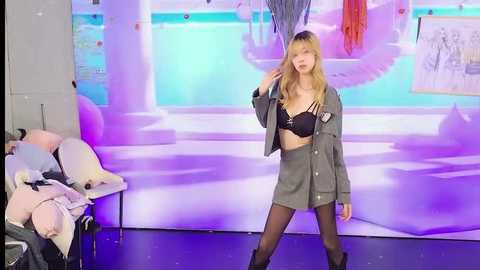 Media: Video of a young woman with long blonde hair in a black lace bra and gray blazer, standing in front of a vibrant, abstract mural with purple lighting, surrounded by white chairs and scattered papers.