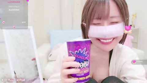 Media: Video of a smiling young Asian woman with straight brown hair, wearing a white mask, a purple mug, and a pink robe, in a bright, clean bedroom.