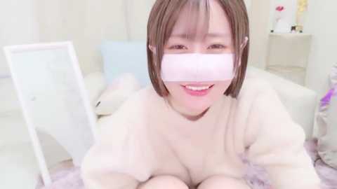 Media: A video of a smiling Asian woman with short brown hair, wearing a white mask and a cream-colored sweater, lying on a white bed with soft, pastel-colored pillows in a bright, minimalistic room.