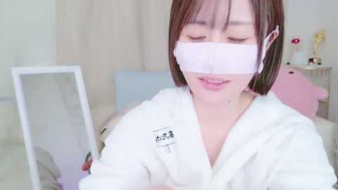 Media: A video of a young Asian woman with straight, shoulder-length brown hair, wearing a white bathrobe and a clear face mask, seated indoors, smiling gently, in a soft, pastel-colored room with light curtains.