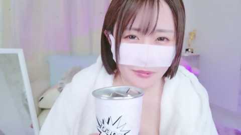 Media: Video of a young woman with shoulder-length brown hair, wearing a white robe and white mask, holding a white mug with a cartoon face design, in a softly lit room with pastel walls and a teddy bear.