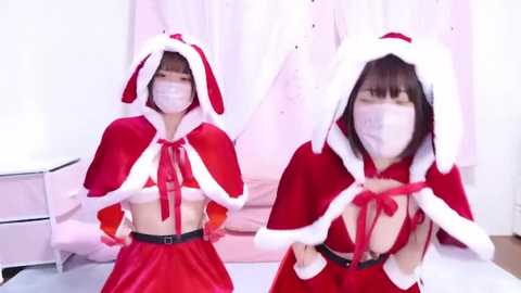 Media: Video of an East Asian woman in a revealing Santa costume, featuring a red velvet cape with white fur trim, matching gloves, and a white face mask. She has a slender physique, fair skin, and short black hair. The background is white with pink accents.