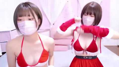 Media: Video of an East Asian woman with short brown hair, wearing a red bikini top and matching skirt, a face mask, and red arm sleeves, in a brightly lit, minimalistic room.