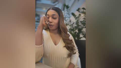 Media: Video of a young Black woman with long, wavy brown hair, wearing a cream ribbed sweater, sitting indoors, smiling with eyes closed, holding a phone to her ear. Background includes green plants and white walls.